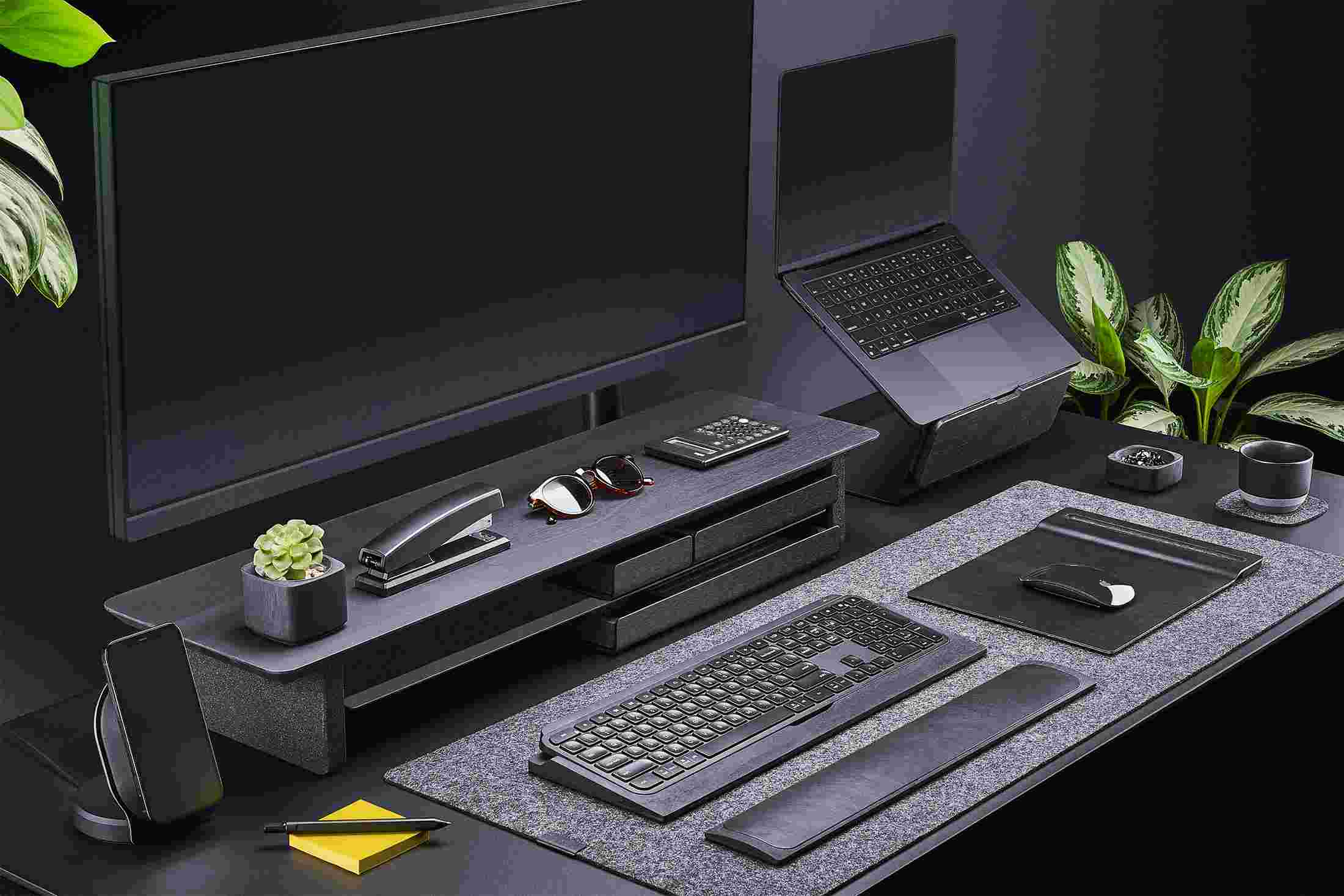 All-black everything—monitor stand, laptop stand, and iPhone stand, on black desk with black felt desk pad.