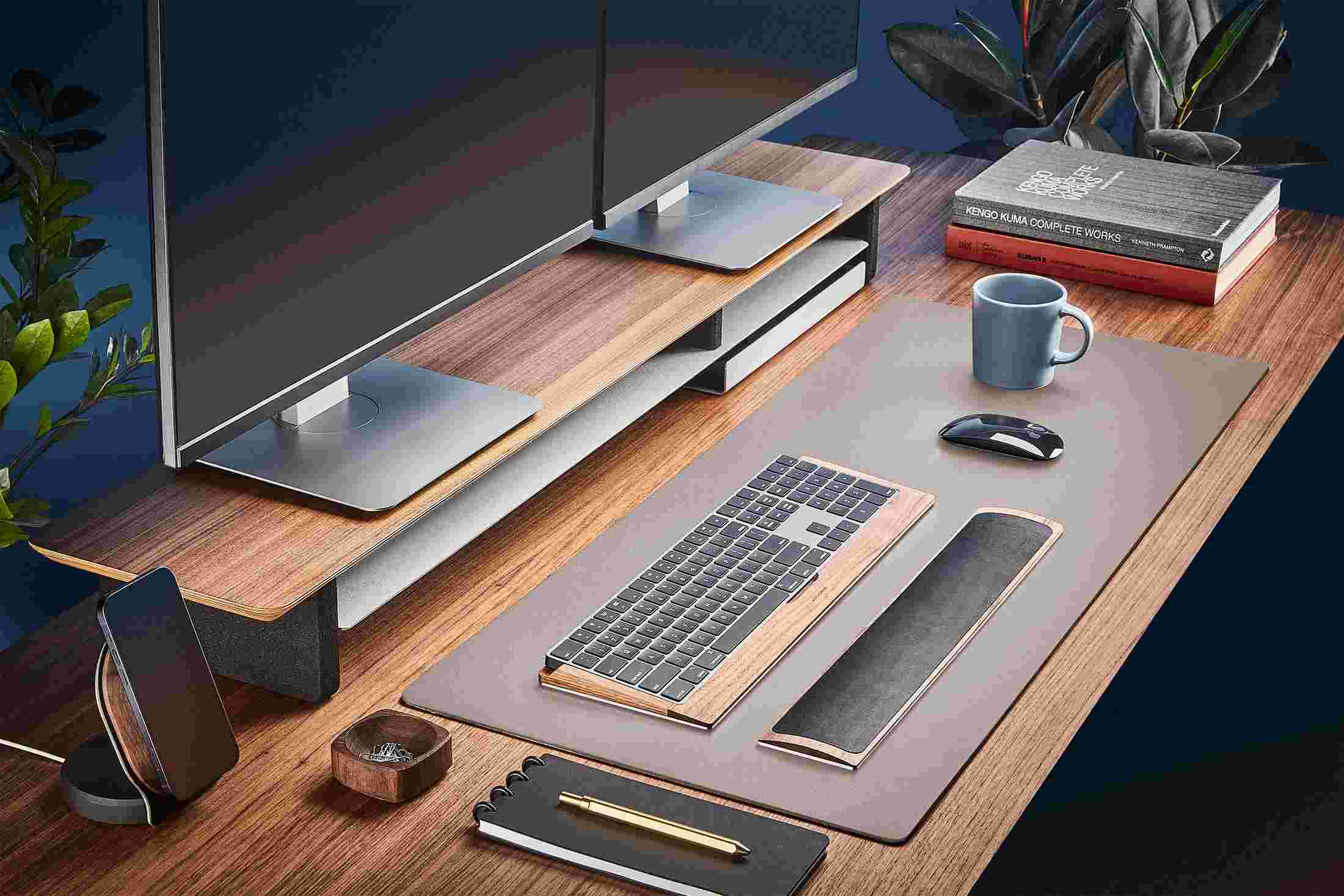 The Medium Matte Desk Pad in Dark Grey runs the entire width of a standard desk, pictured here with walnut wood double-wide monitor stand.