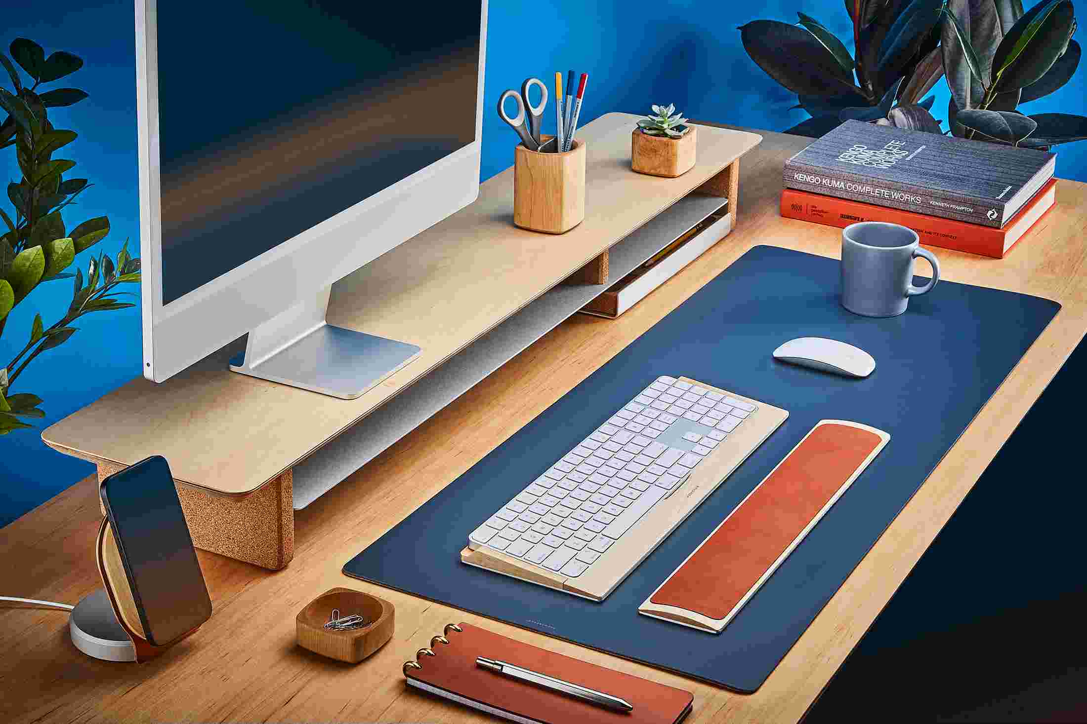 The Medium Matte Desk Pad in Navy Blue runs the entire width of a standard desk, pictured here with maple wood double-wide monitor stand.