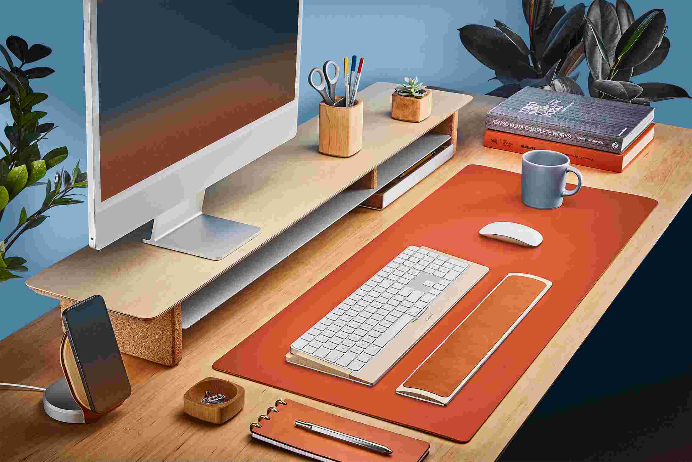 The Medium Matte Desk Pad in Orange runs the entire width of a standard desk, pictured here with matte black double-wide monitor stand.
