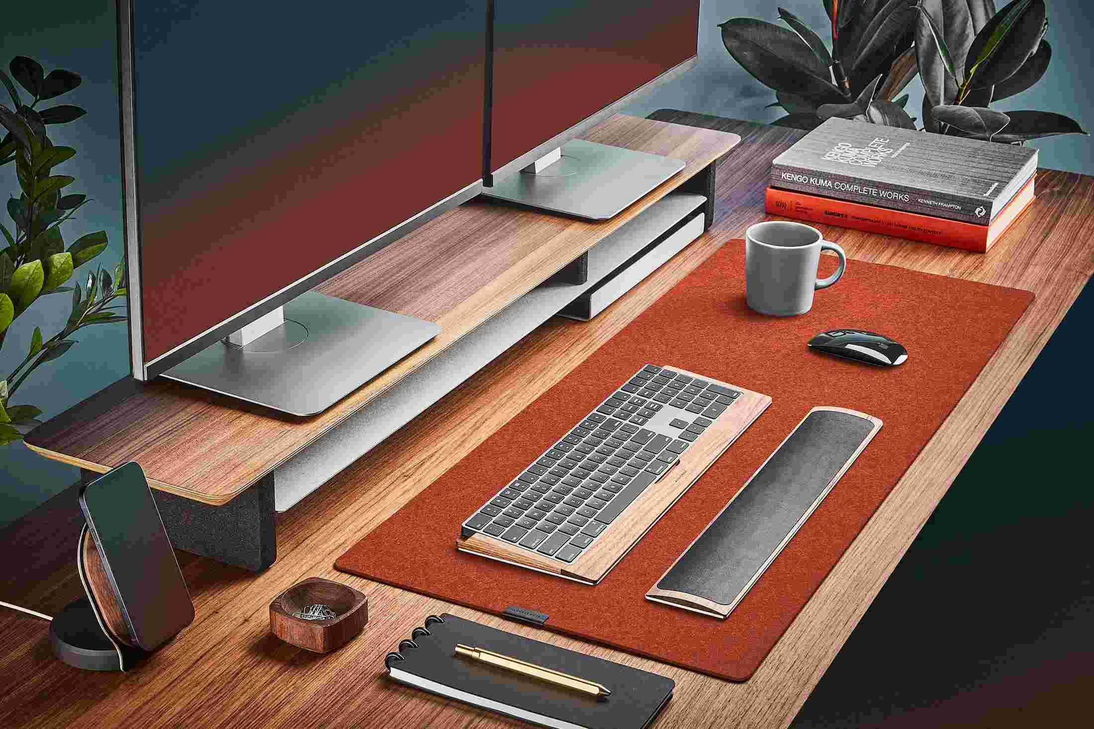 The Dark Orange Medium Plus Wool Felt Desk Pad runs the entire width of a standard desk, pictured here with walnut wood double-wide monitor stand.